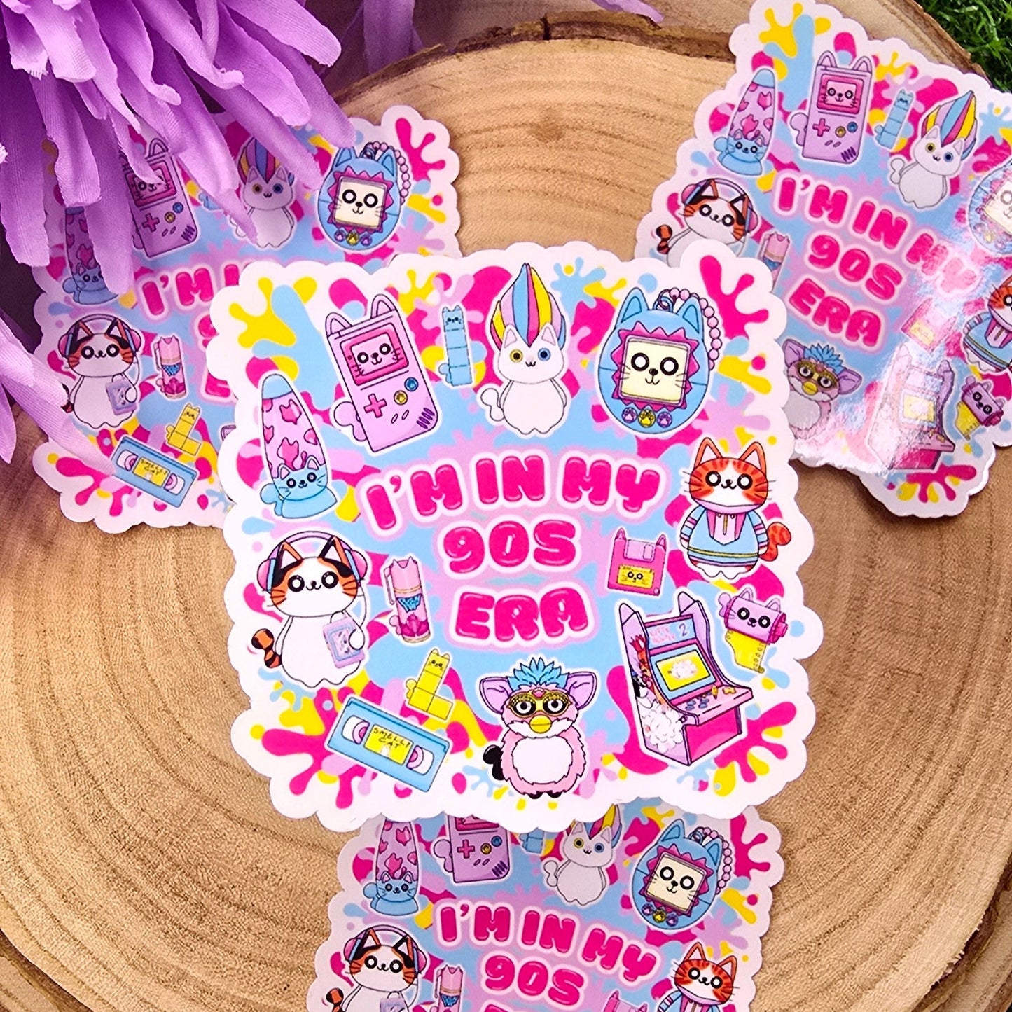 A colorful, die-cut sticker is displayed on a round wooden slab, which rests on vibrant green grass. Behind the wood slab are vivid purple flowers with soft petals. The sticker features a playful, 1990s theme with cute cat characters and nostalgic icons such as a Tamagotchi, video tape, arcade machine, and a character resembling a Furby. In bold pink text in the center, it reads: "I'M IN MY 90s ERA." The background of the sticker has splashes in yellow, blue, & purple adding to the retro aesthetic.