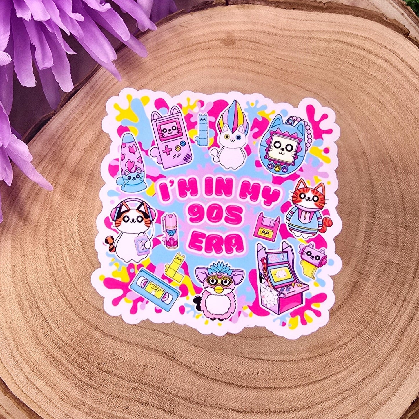 A colorful, die-cut sticker is displayed on a round wooden slab, which rests on vibrant green grass. Behind the wood slab are vivid purple flowers with soft petals. The sticker features a playful, 1990s theme with cute cat characters and nostalgic icons such as a Tamagotchi, video tape, arcade machine, and a character resembling a Furby. In bold pink text in the center, it reads: "I'M IN MY 90s ERA." The background of the sticker has splashes in yellow, blue, & purple adding to the retro aesthetic.