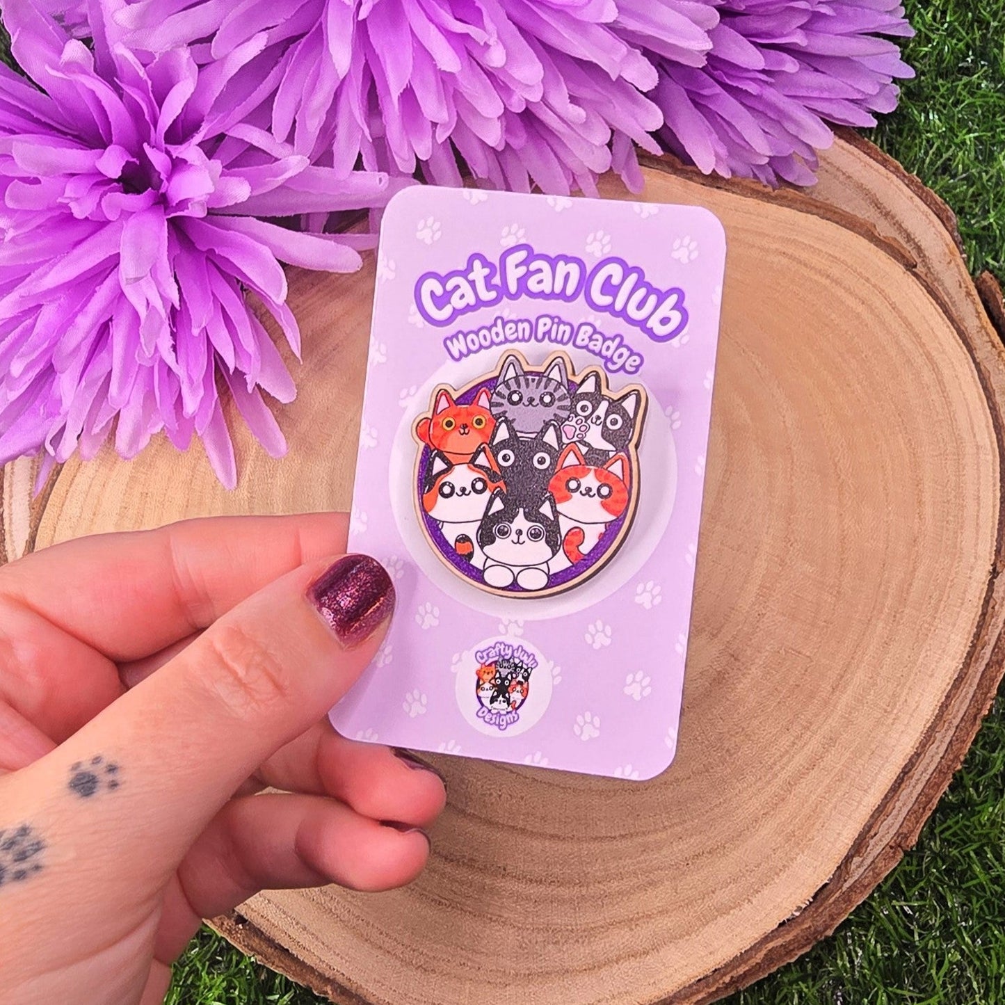 Hand holding a 'Cat-Fan Club' badge with purple flowers in the background