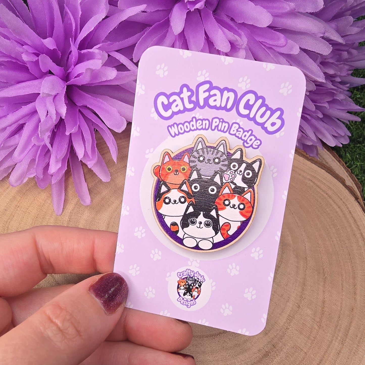 Cat-themed wooden pin badge on a card held by a hand with purple flowers in the background