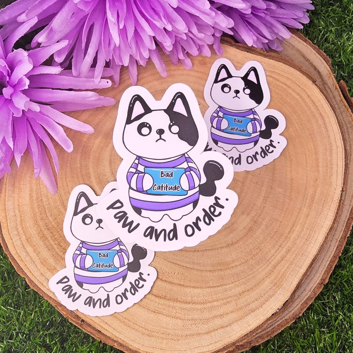 Three cat-shaped stickers with text on a wooden surface with purple flowers in the background.