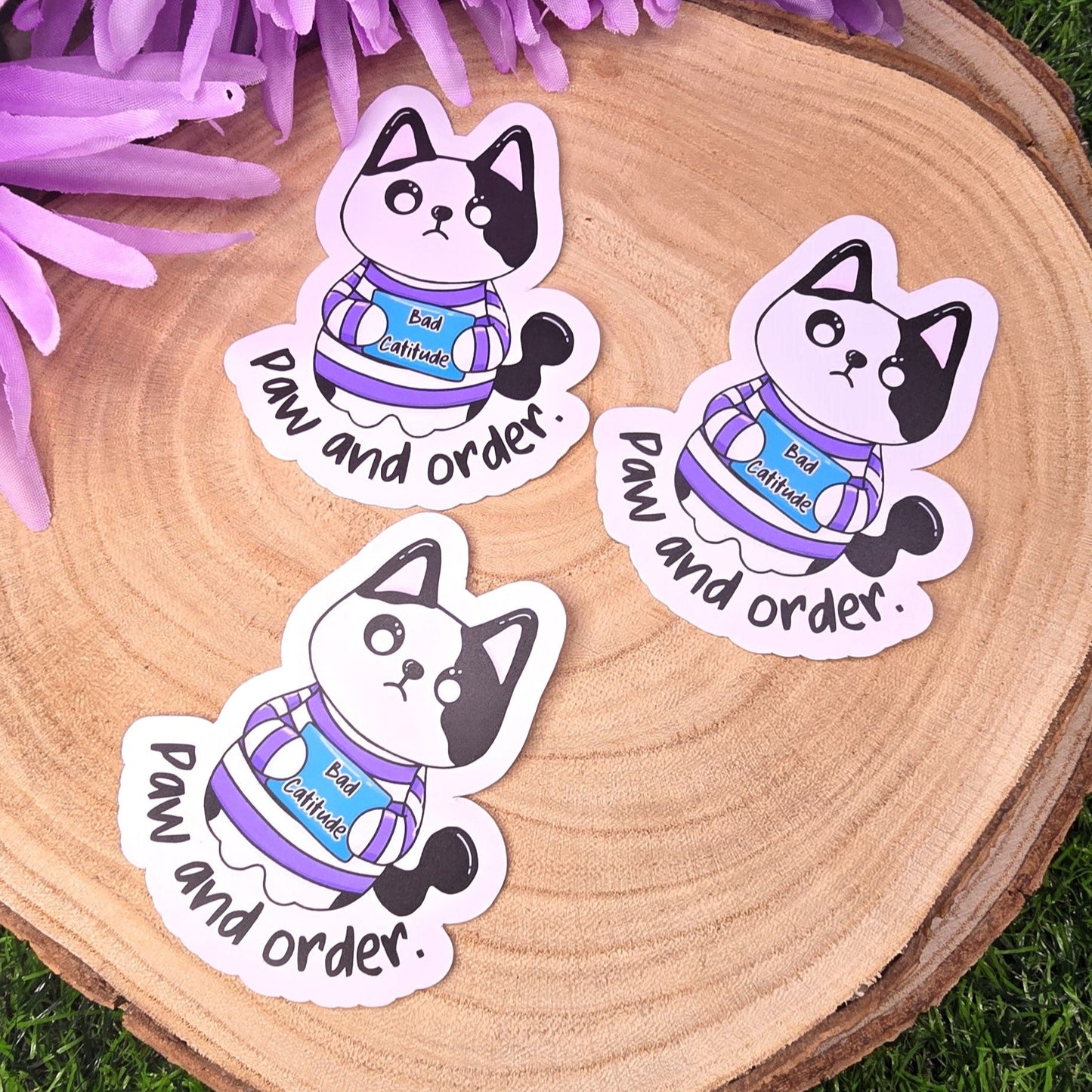 Three stickers of a cat in a sweater with text on a wooden surface surrounded by purple flowers.