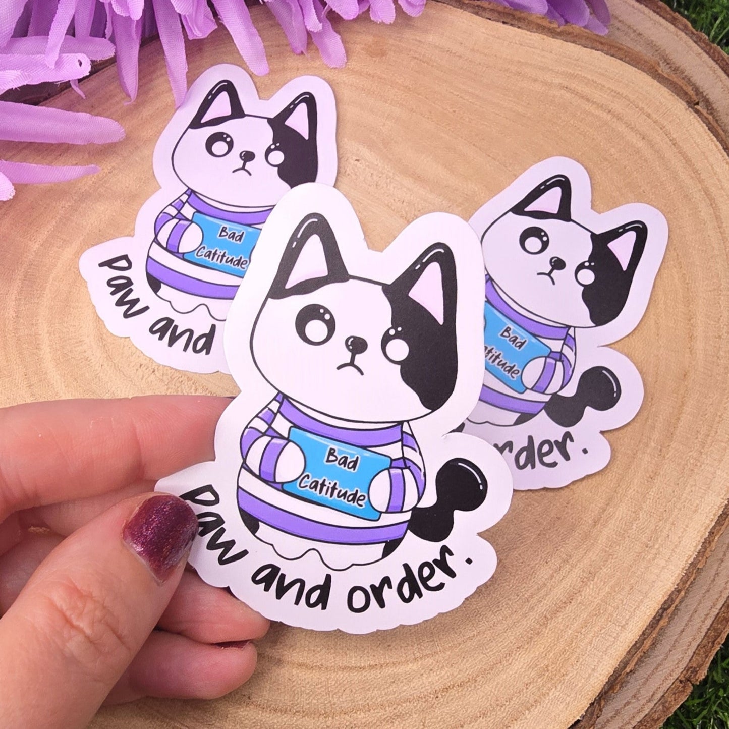 Three cute cat-shaped stickers with text on a wooden surface with purple flowers in the background.