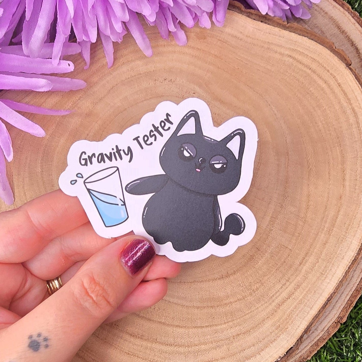 Sticker of a cat labeled 'Gravity Tester' held over a wooden surface with purple flowers in the background.