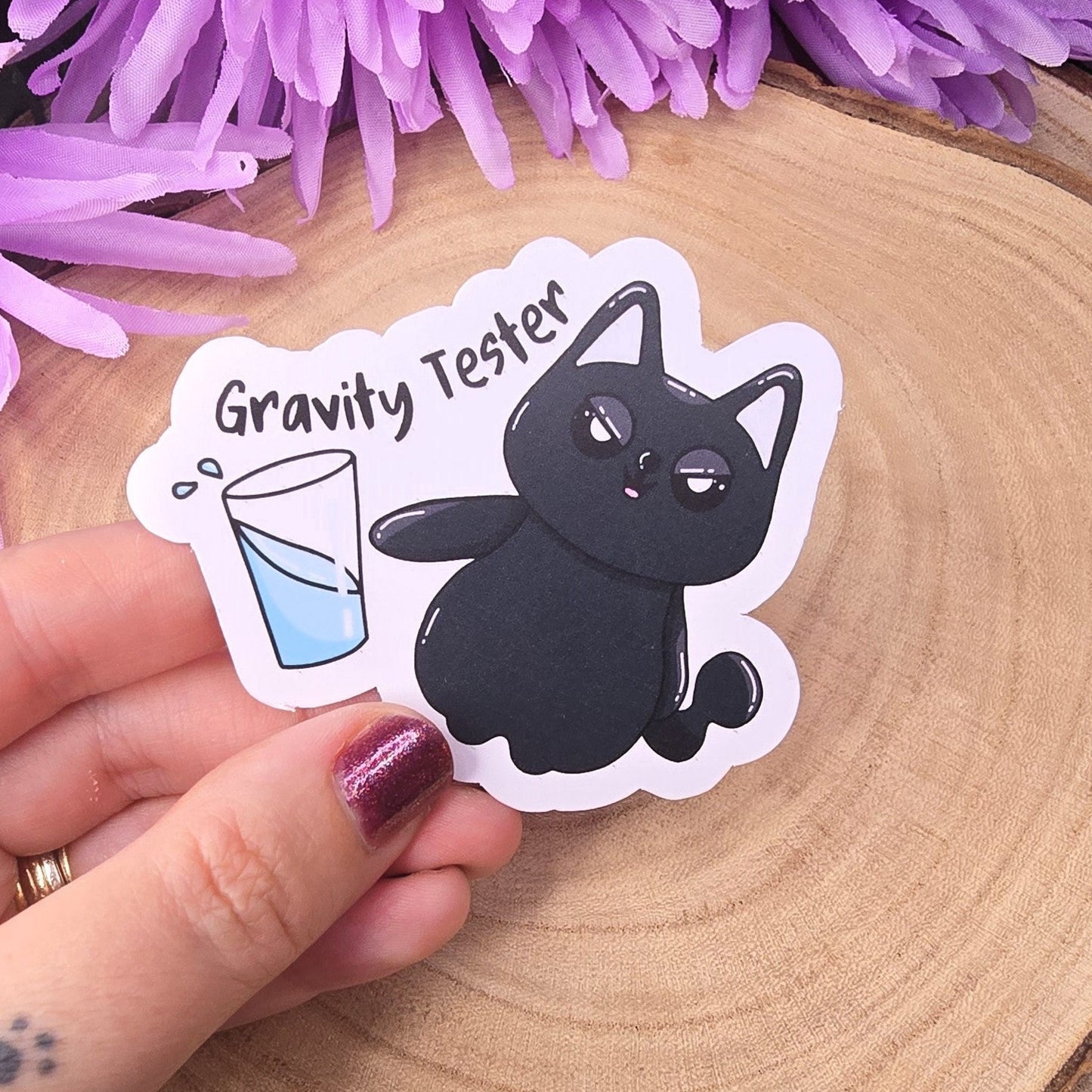 Sticker of a black cat labeled 'Gravity Tester' held over a wooden surface with purple flowers in the background.