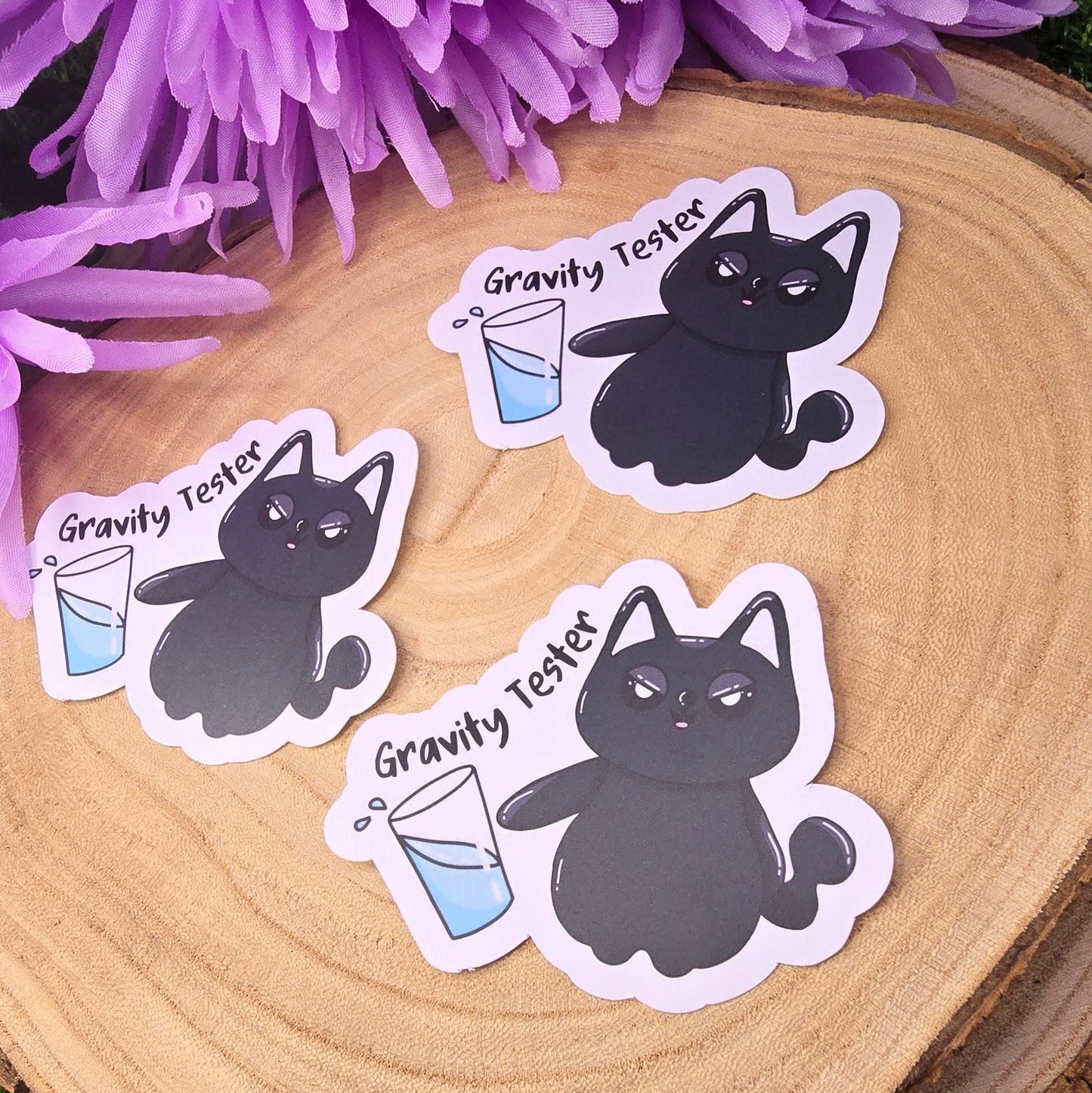 Three black cat stickers with 'Gravity Tester' text on a wooden surface with purple flowers in the background.