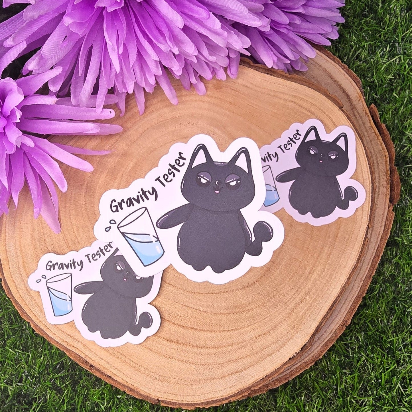 Stickers of a cat character with 'Gravity Tester' text on a wooden surface with purple flowers.