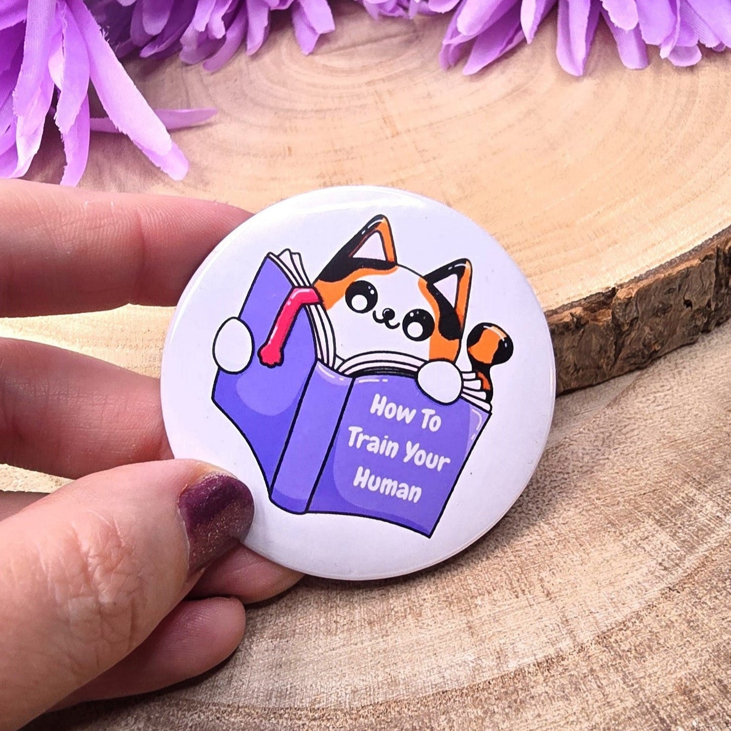 Button with a cat illustration and text held by a hand against purple flowers on a wooden surface