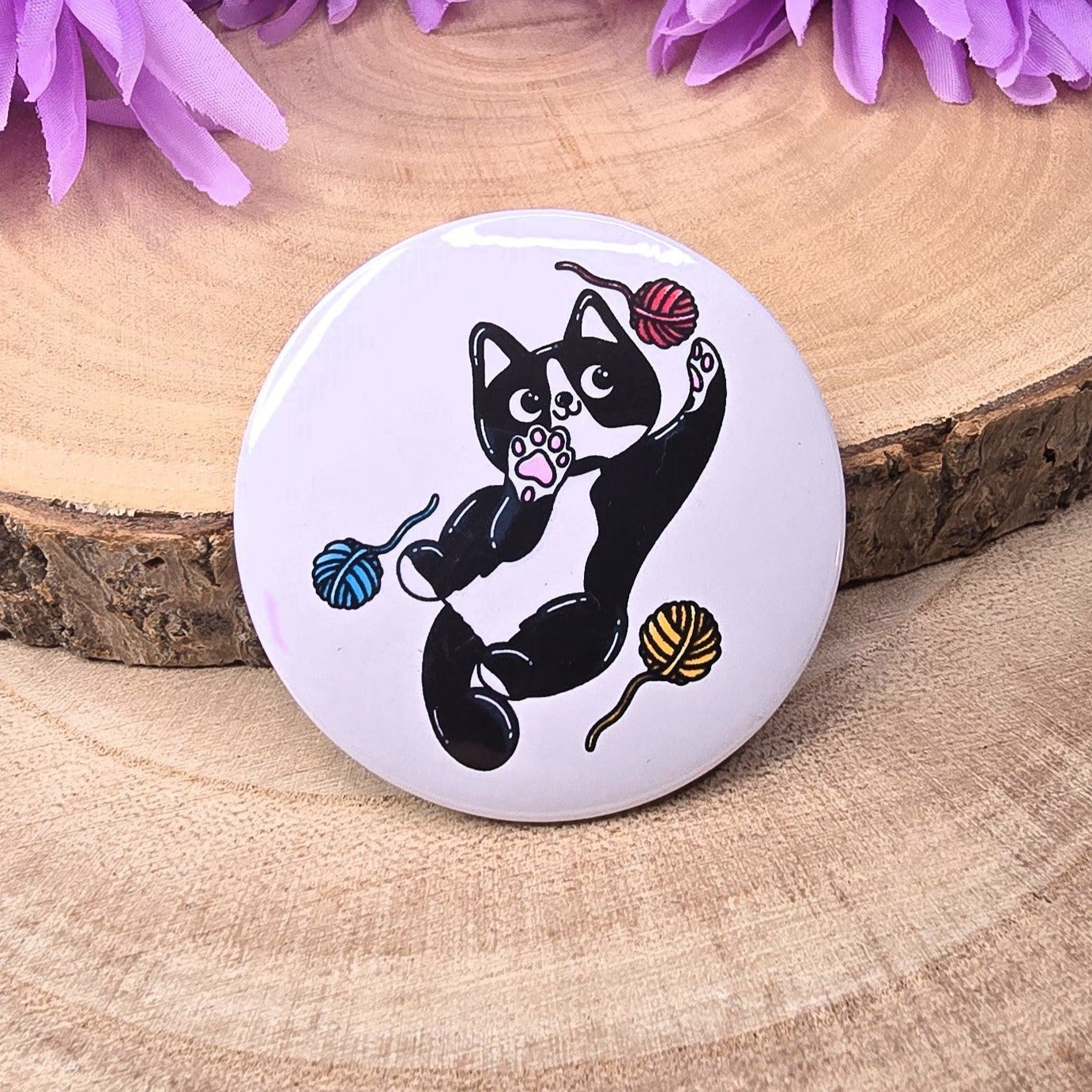 Button badge with a black cat design on a wooden surface with purple flowers