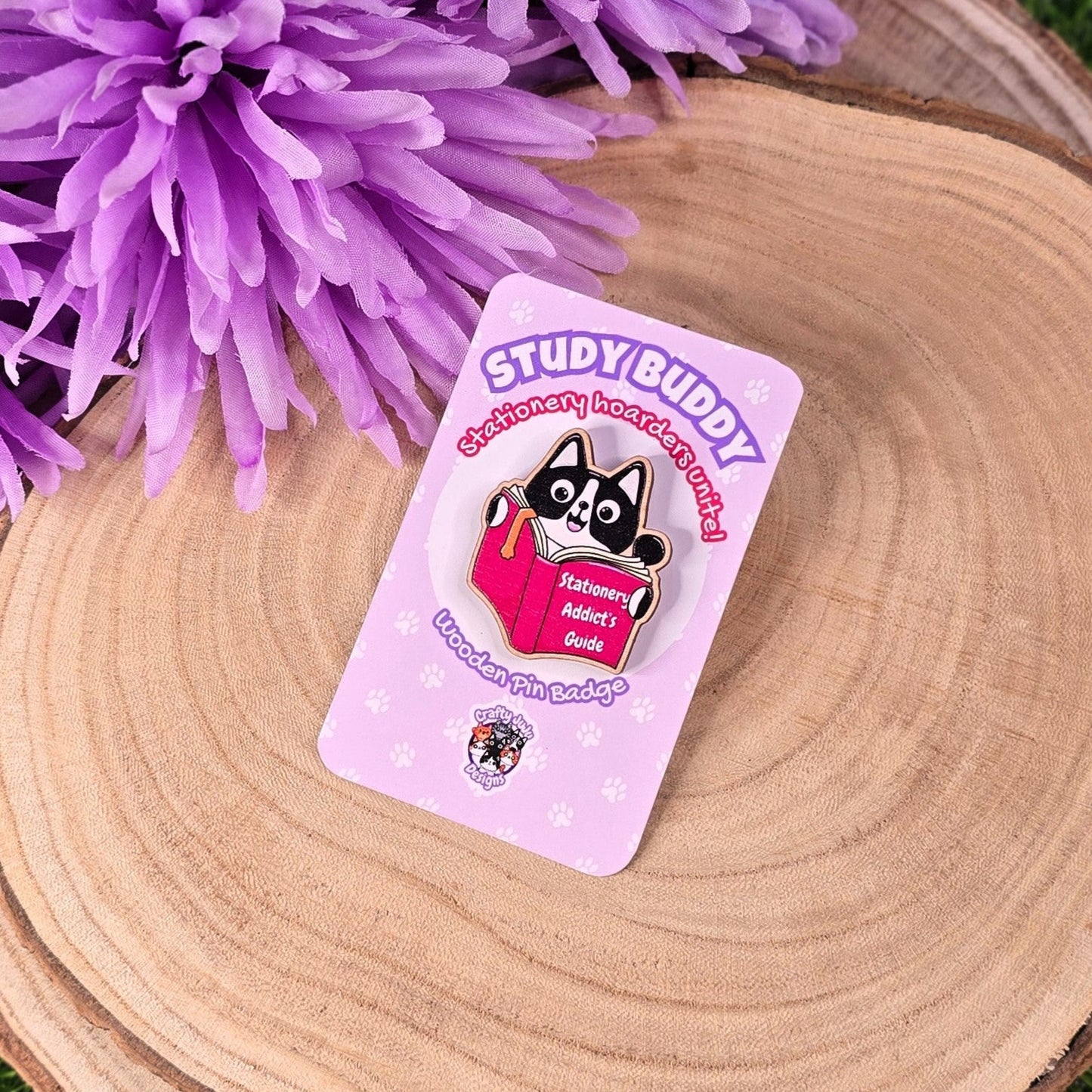Pin with cartoon character on a pink card on a wooden surface with purple flowers in the background