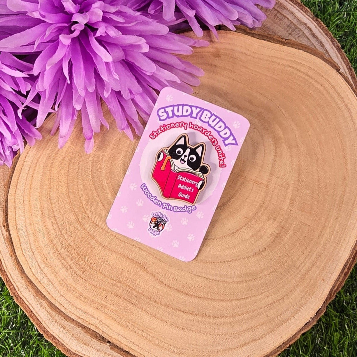 Pin with 'Study Buddy' design on a pink card, placed on a wooden surface with purple flowers in the background.
