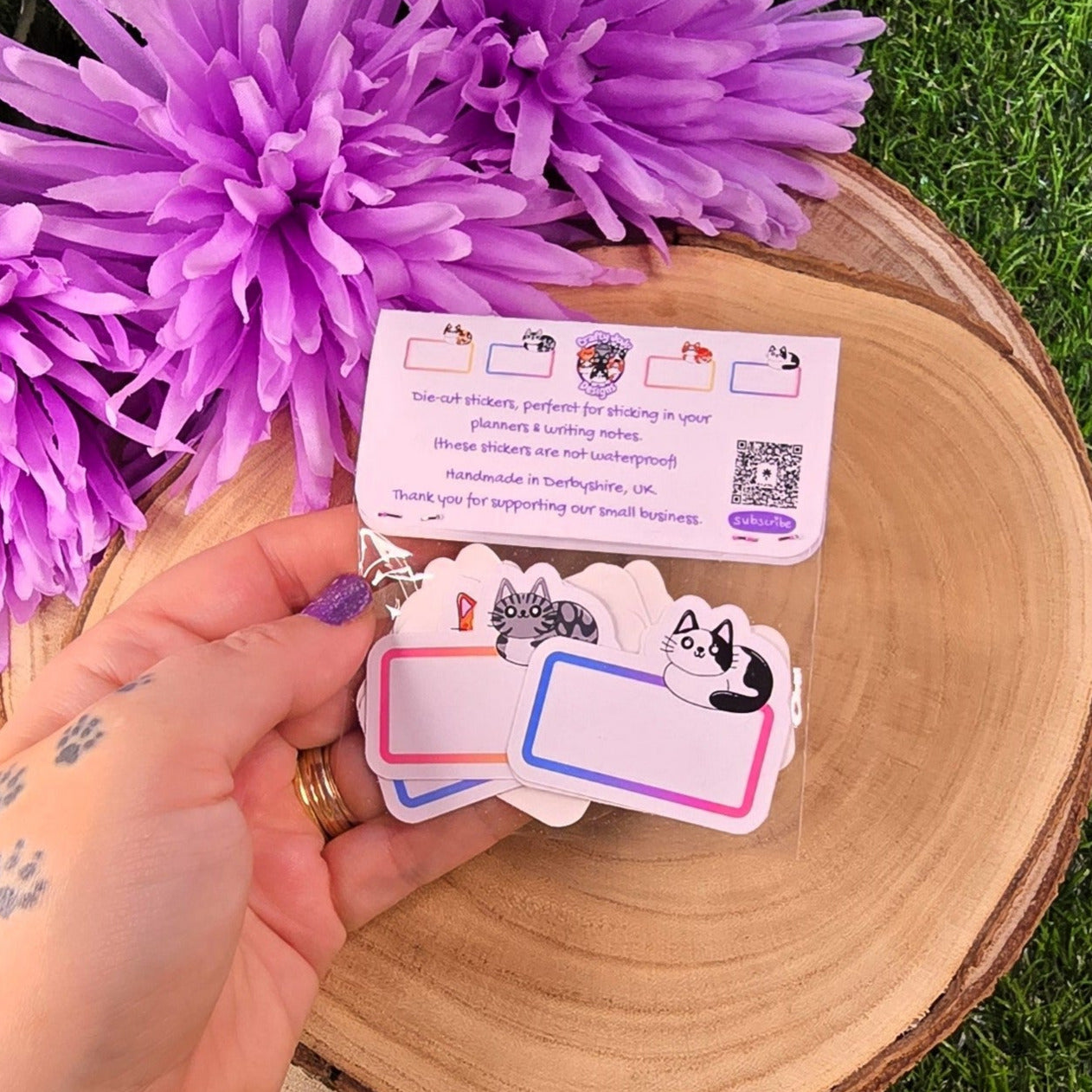 Hand holding a set of cat stickers with purple flowers and a wooden board in the background.