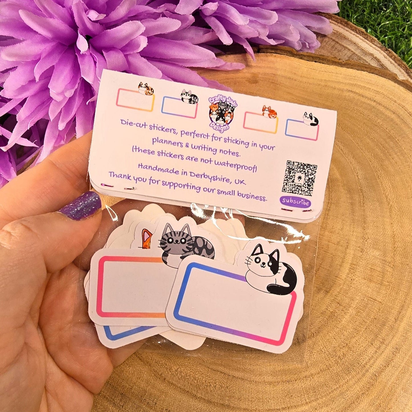 Hand holding a set of die-cut stickers with cat illustrations, placed on a wooden surface with purple flowers in the background.