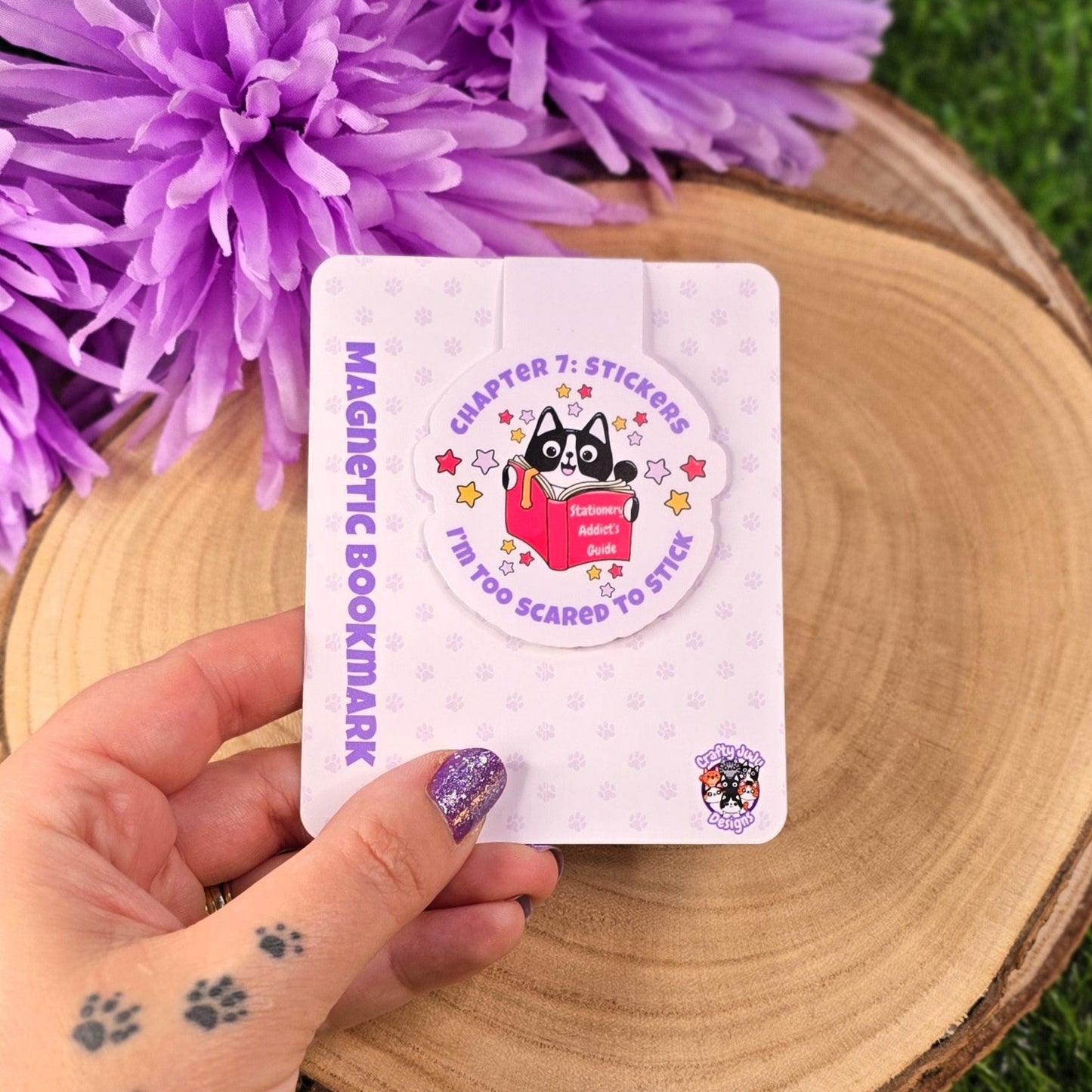 Magnetic bookmark with a cat design held by a hand with paw print tattoos, against a background of purple flowers on a wooden block.