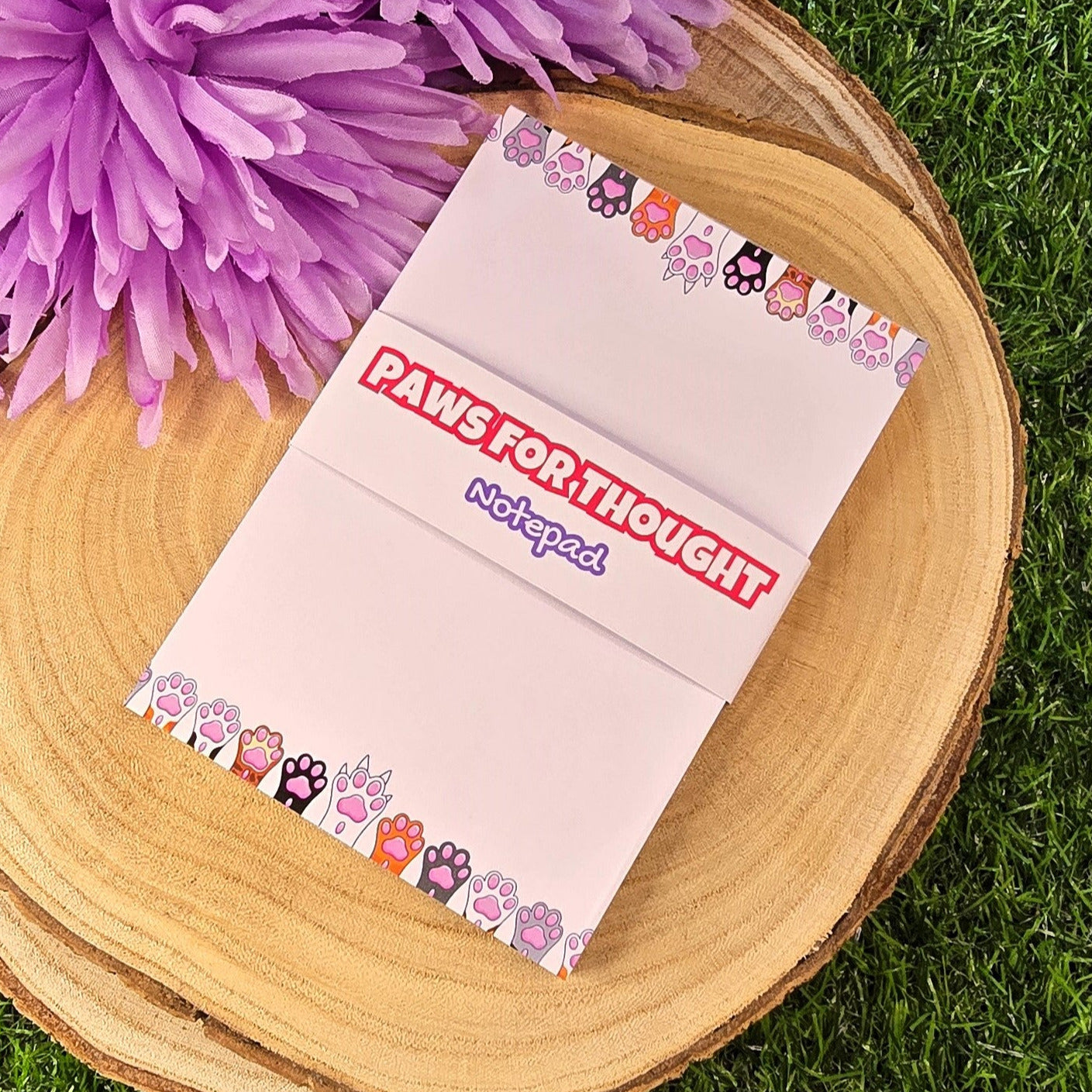 Notebook titled 'Paws for Thought' on a wooden surface with purple flowers and grass in the background