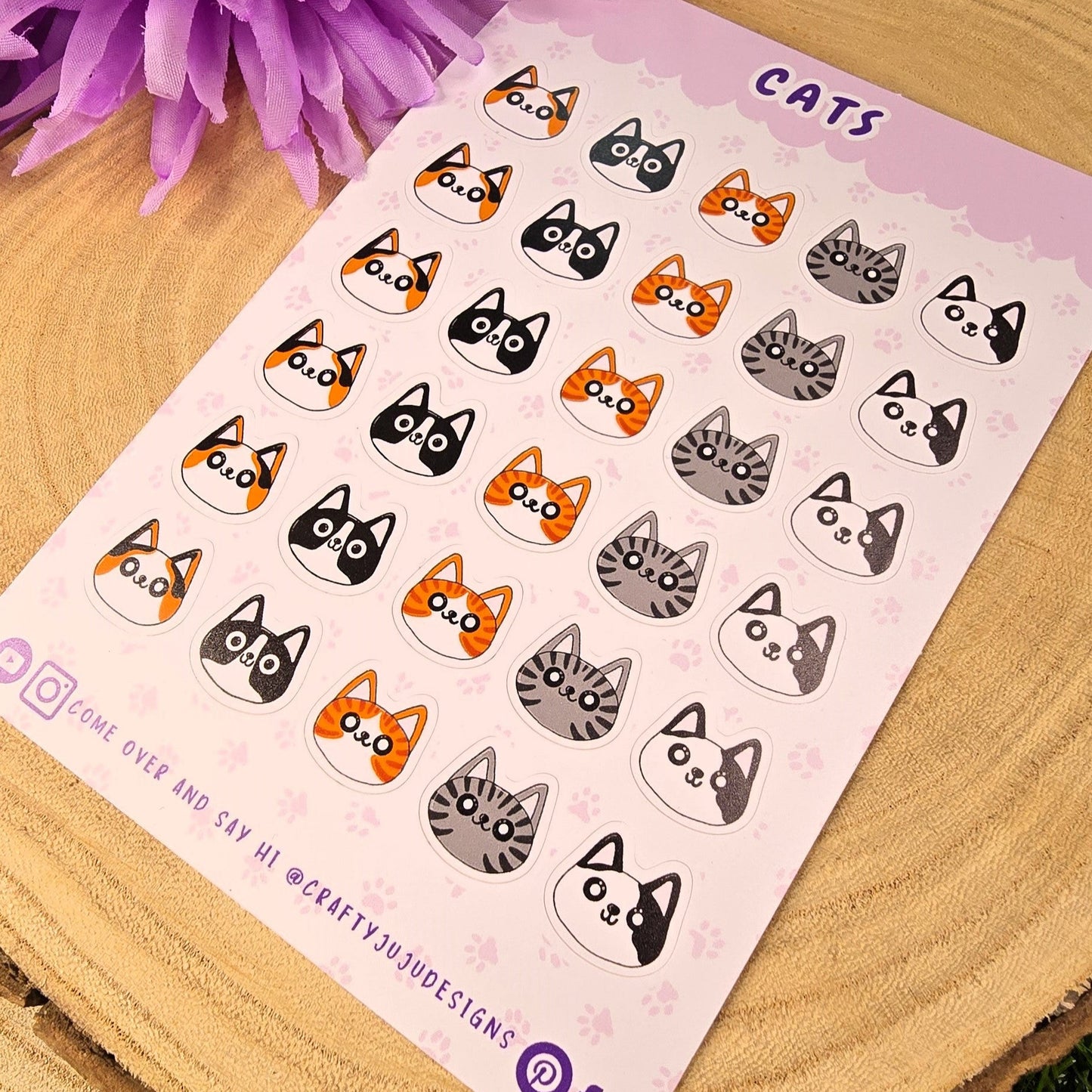 Sheet of cat-themed stickers on a wooden surface with purple flowers in the background