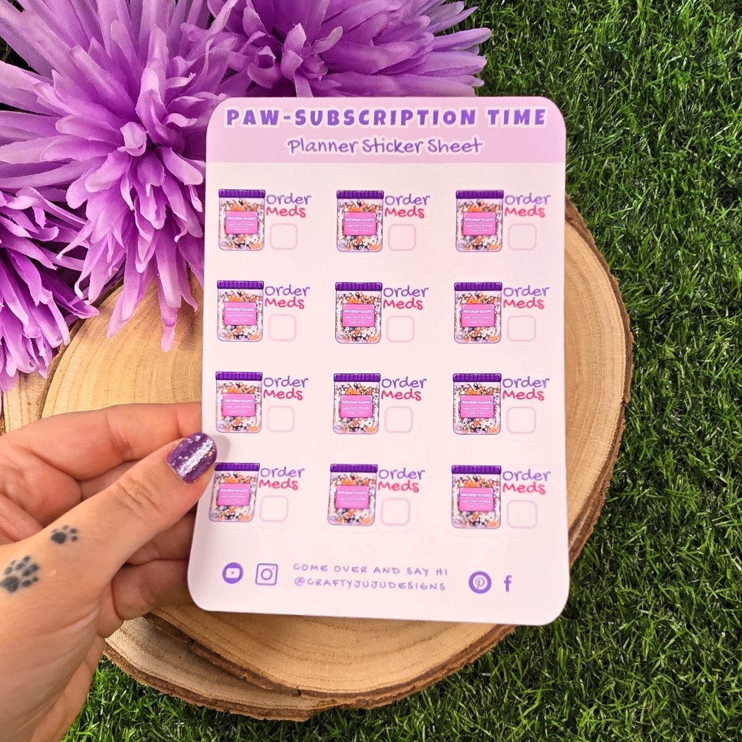 Hand holding a planner sticker sheet 'Paw-Subscription Time' stickers on grass with purple flowers.