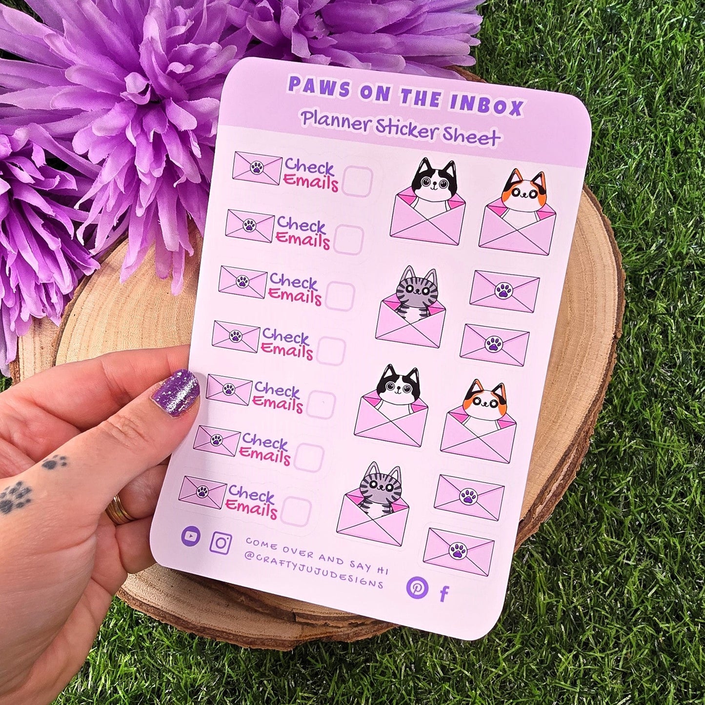 Sticker sheet with cat-themed designs held by a hand on grass with purple flowers.
