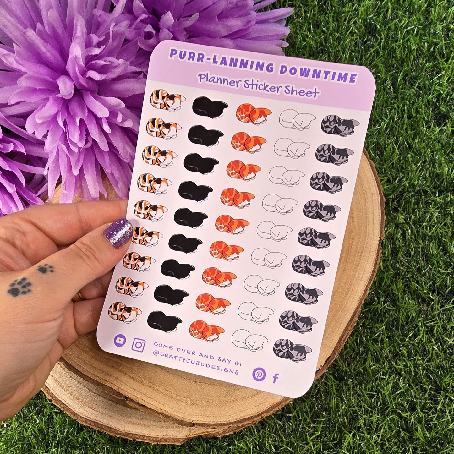 Sheet of planner stickers with cat designs held by a hand on grass with purple flowers.