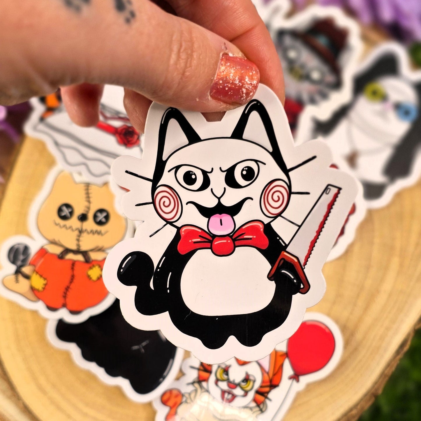Hand holding a sticker of a cat with a bow tie and knife, surrounded by other stickers on a wooden surface.