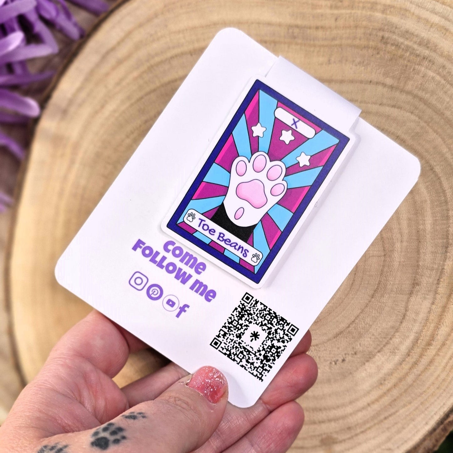 Hand holding a card with a paw print design and QR code, surrounded by purple flowers on a wooden surface.
