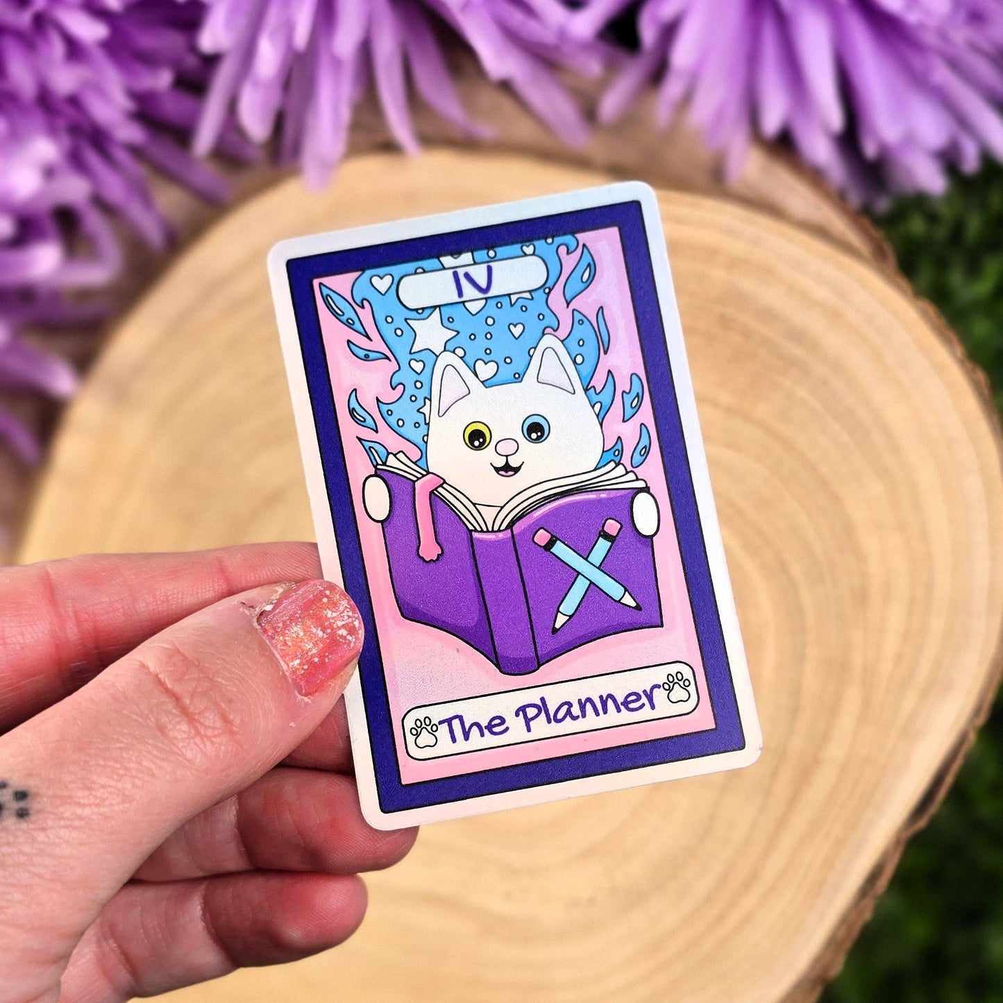 Hand holding a sticker with a cartoon cat design and 'The Planner' text, against a purple flower background.