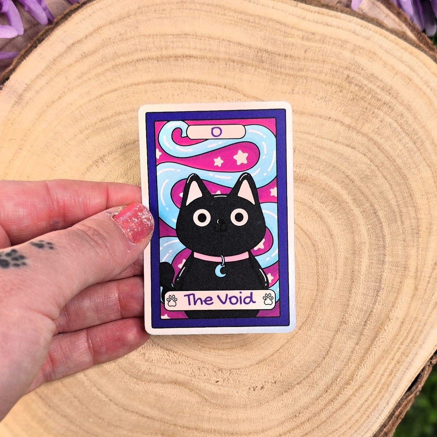 sticker with a black cat illustration labeled 'The Void' held by a hand on a wooden surface with purple flowers.
