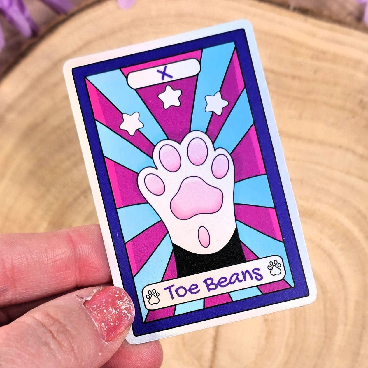 Colorful sticker with a paw print design held by a hand on a wooden surface with purple flowers in the background.