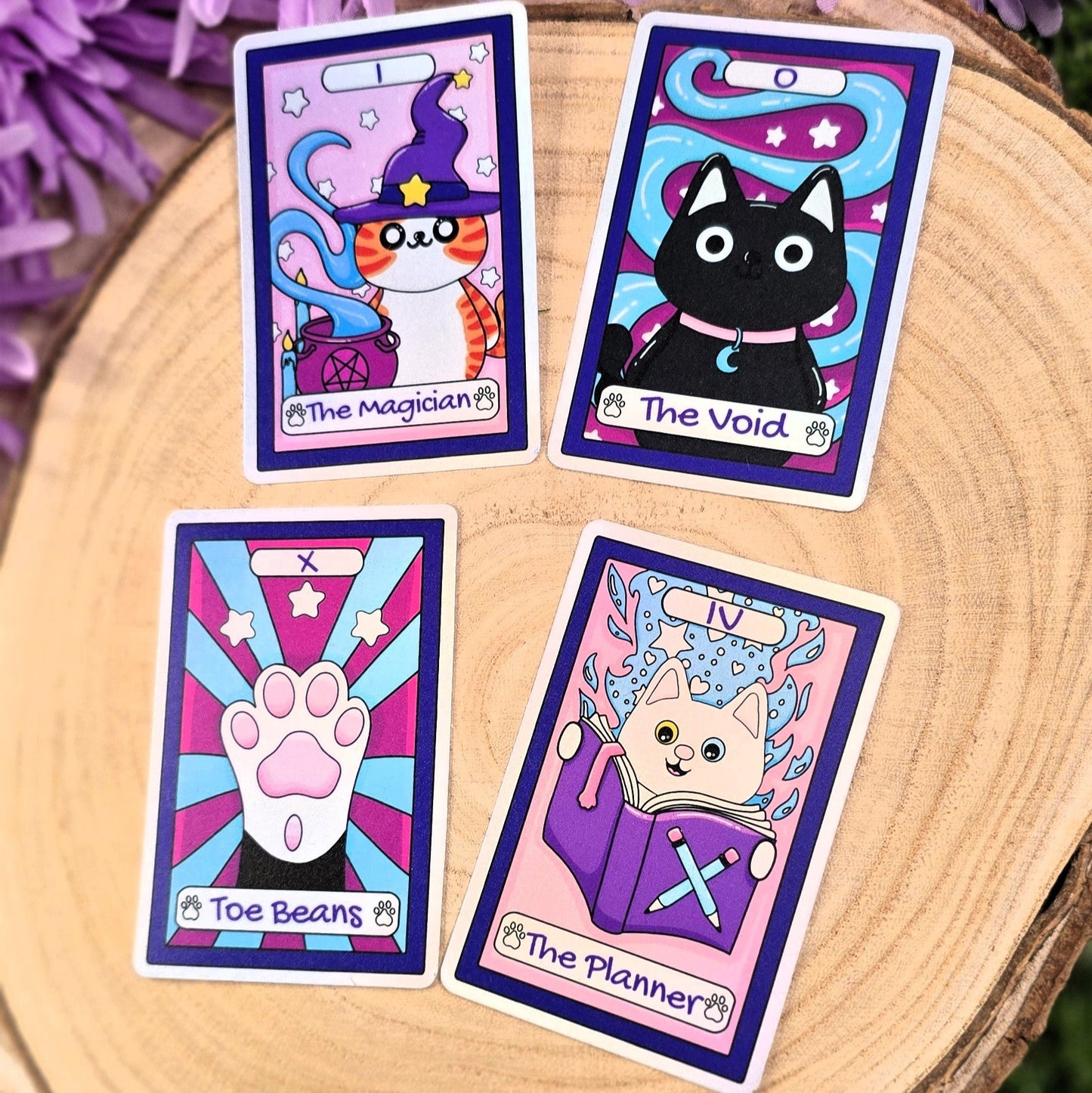 Four tarot card-style stickers with cat illustrations on a wooden surface with purple flowers in the background.