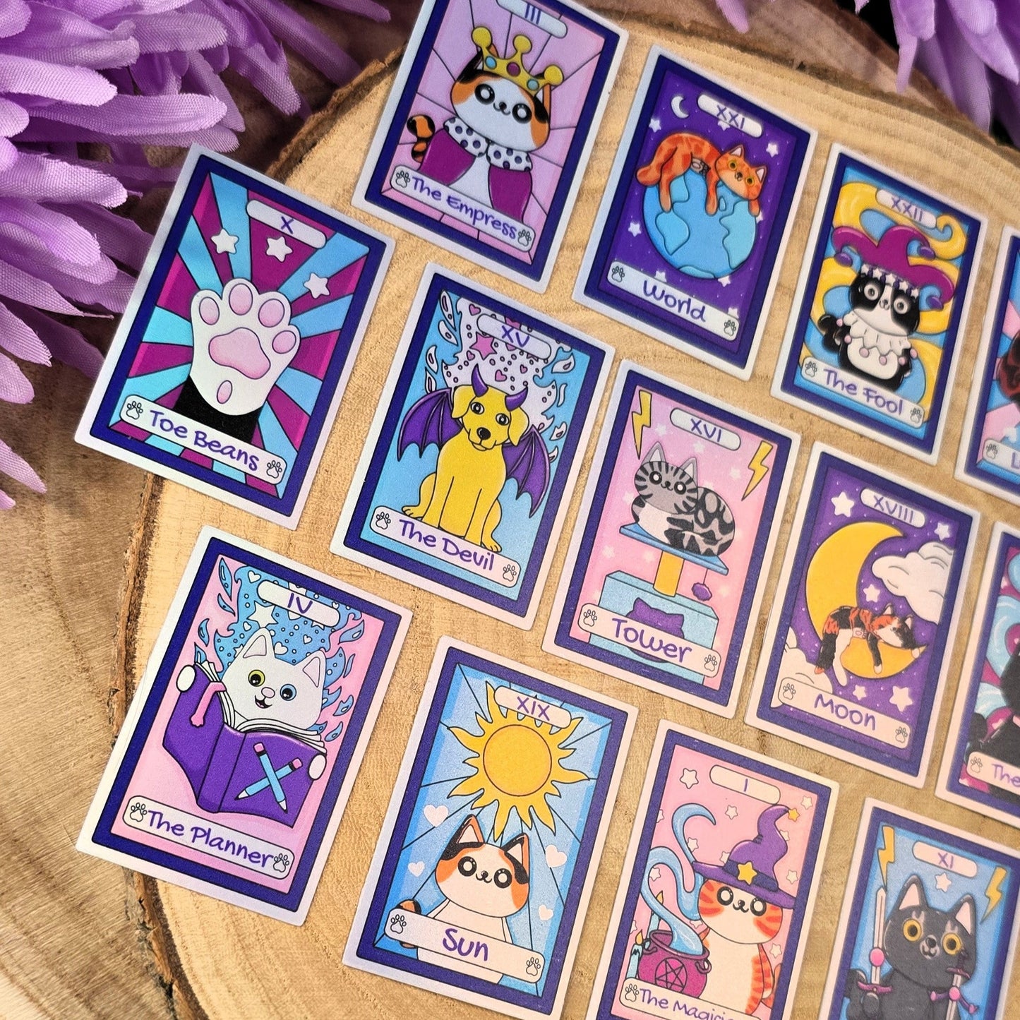 Set of colorful tarot card stickers with cat illustrations on a wooden surface with purple flowers in the background.