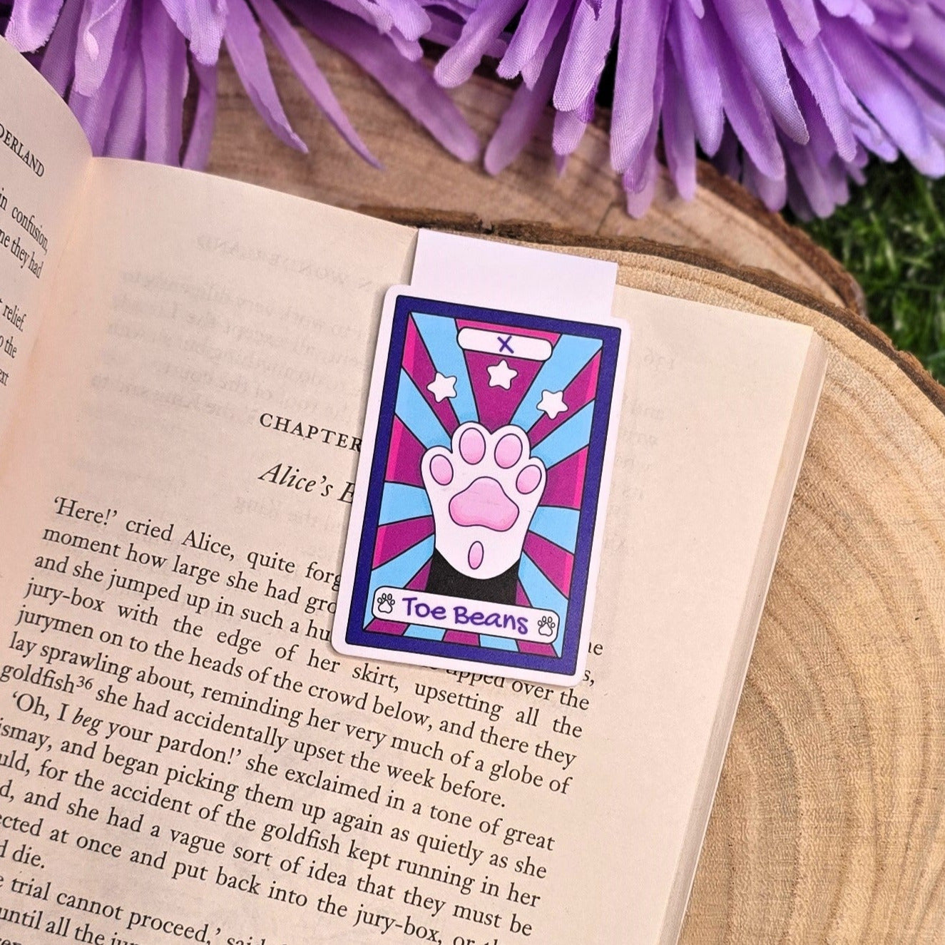 Open book with a colorful bookmark featuring a paw print design, surrounded by purple flowers on a wooden surface.