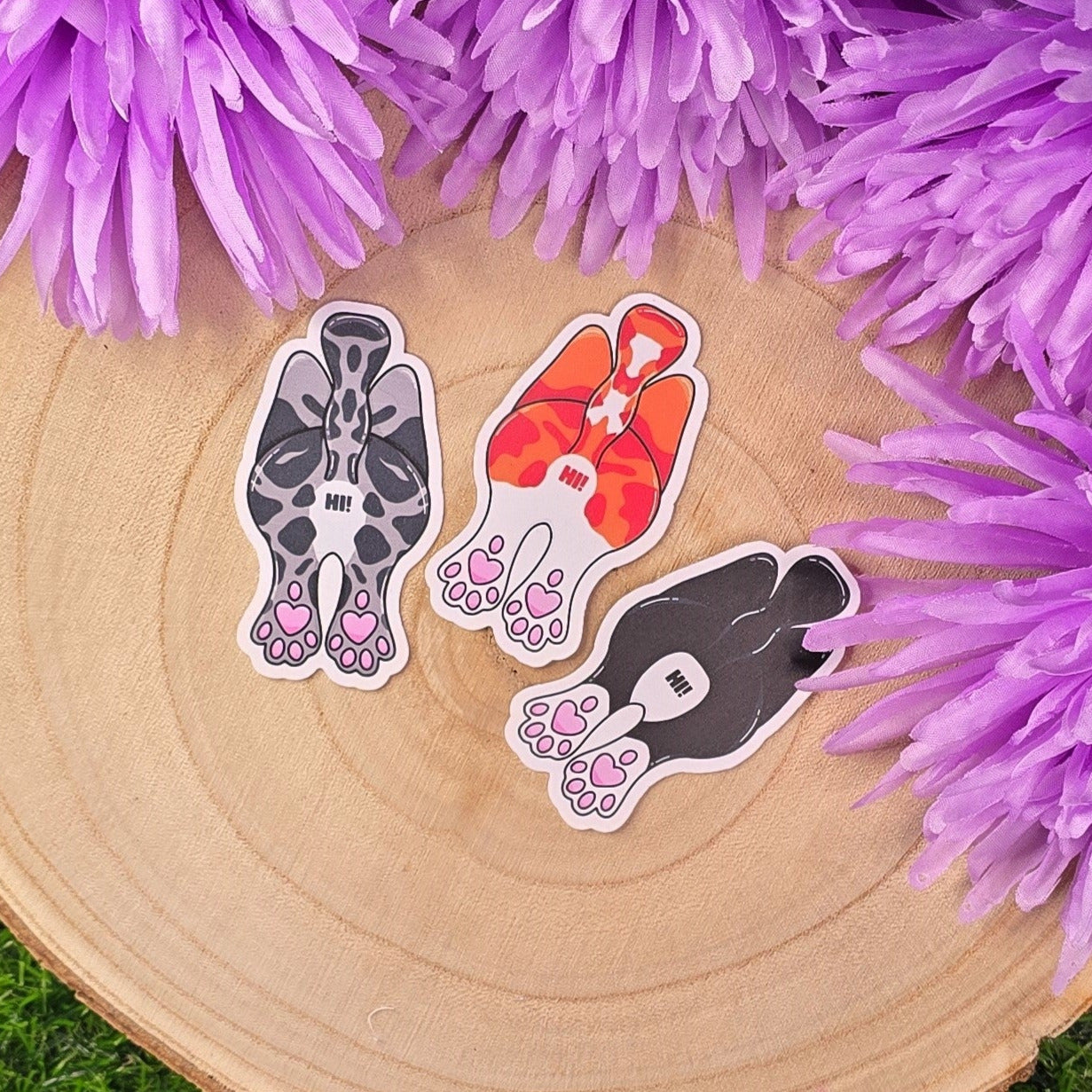 Three cat-shaped stickers on a wooden surface with purple flowers in the background.