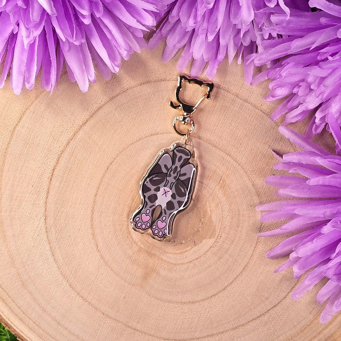 Cat-butt keychain on a wooden surface surrounded by purple flowers
