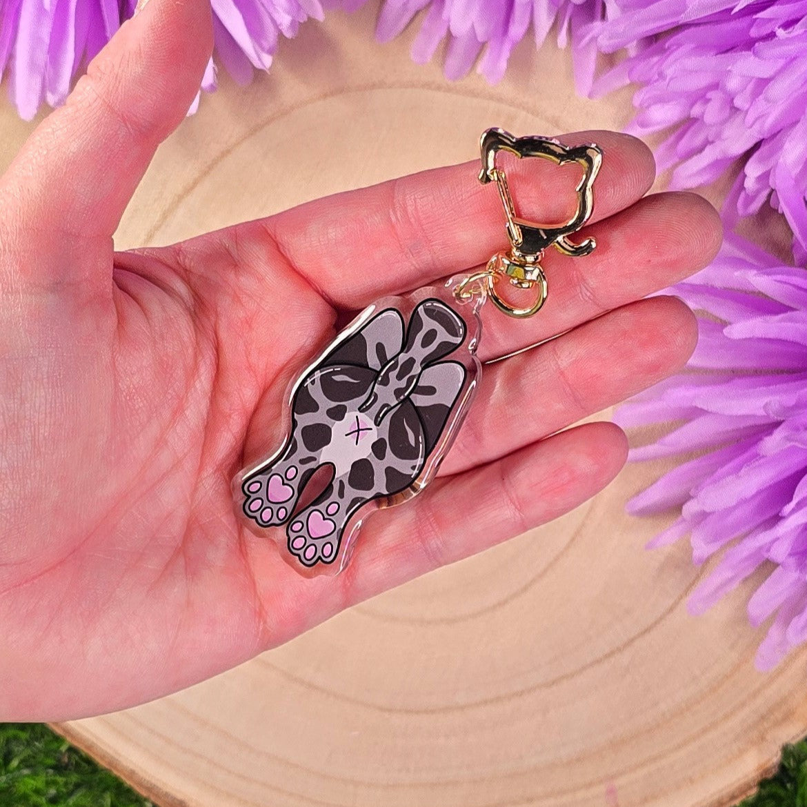 Hand holding a keychain with a cat butt design against a purple flower background