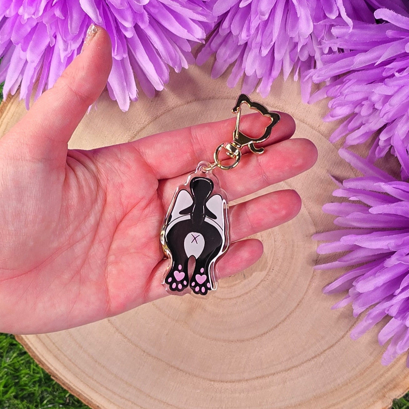 Hand holding a keychain with a black and white cat butt design against a purple floral background