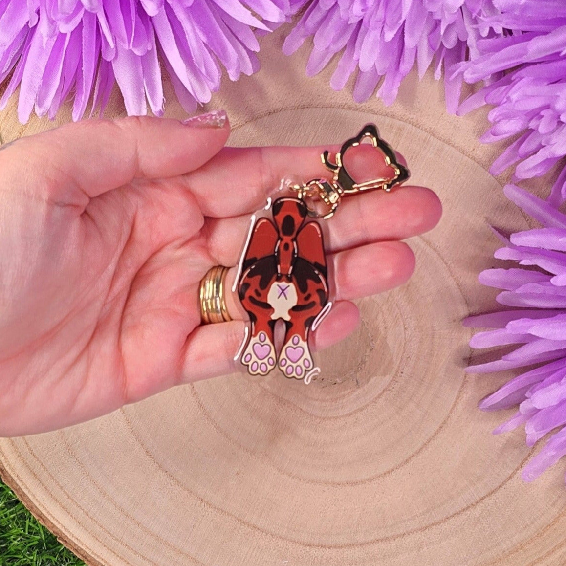 Hand holding a keychain with a design of a brown cat in front of purple flowers on a wooden surface.