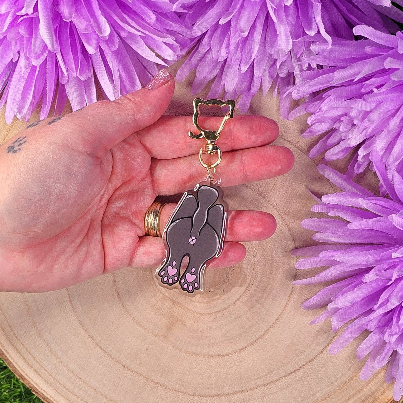 Hand holding a keychain with cat butt design against a background of purple flowers