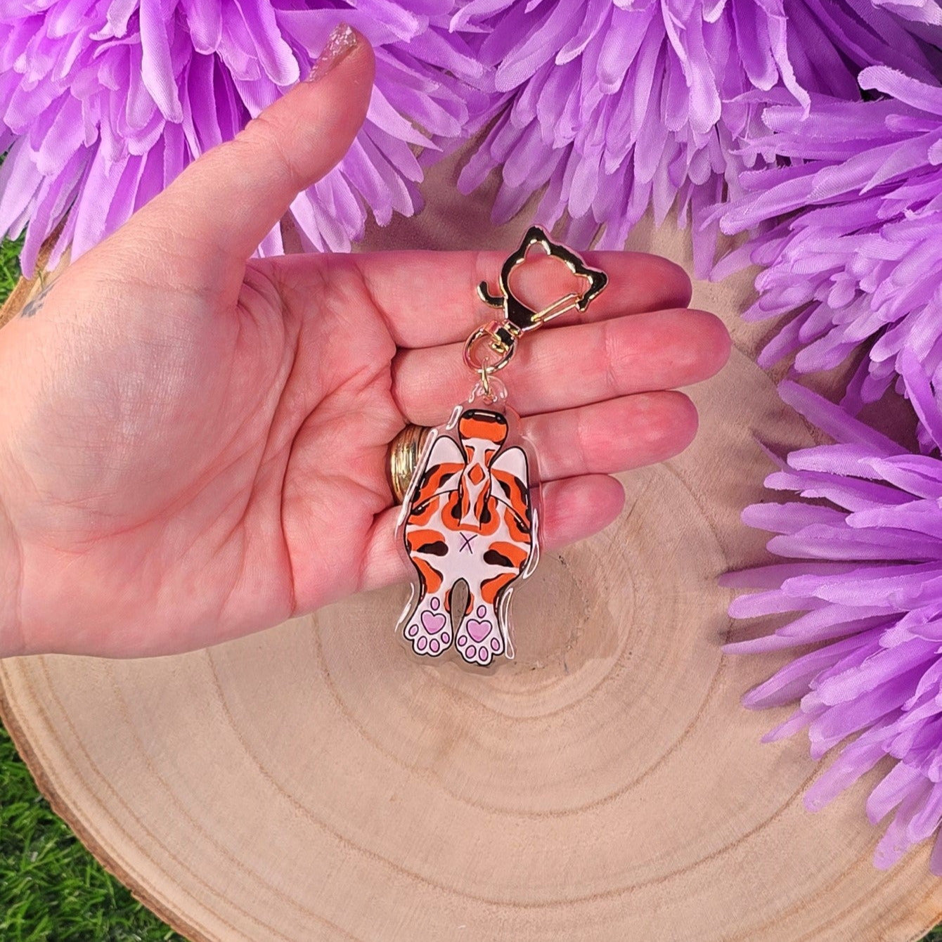 Hand holding a cat butt keychain with purple flowers in the background