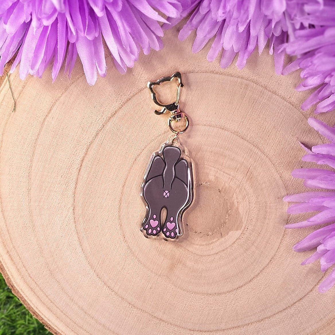 Cat-butt shaped keychain with purple accents on a wooden surface with purple flowers.