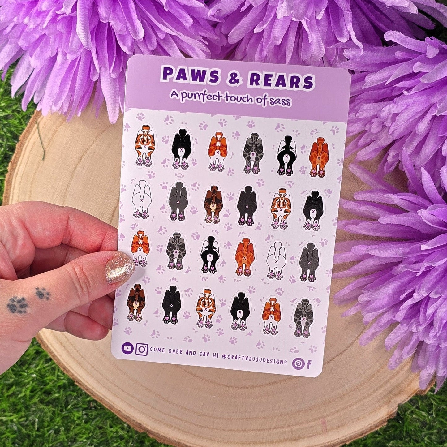 Sheet of stickers with various cat poses held by a hand, surrounded by purple flowers on a wooden surface.