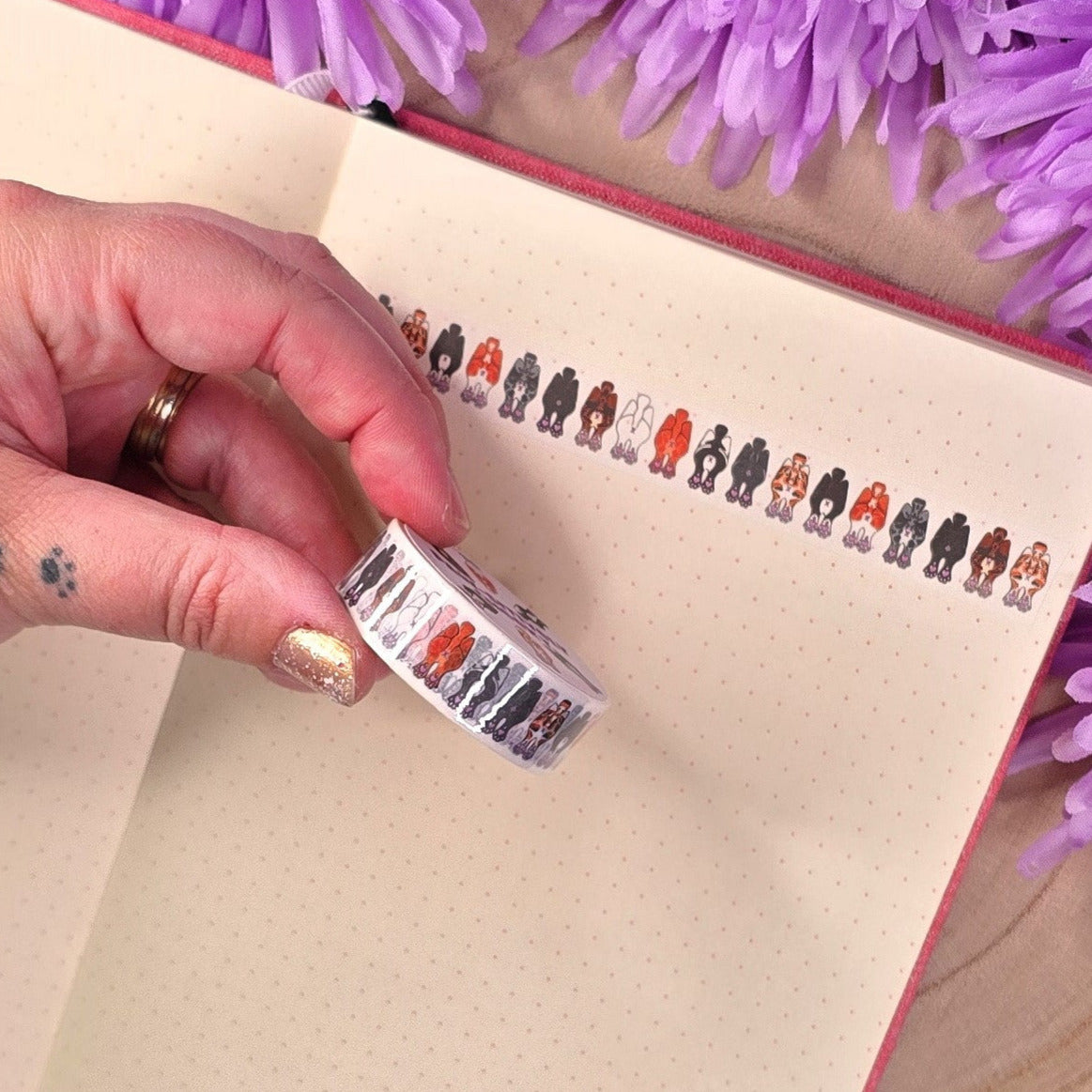 Hand holding a roll of decorative tape with some on a notebook with purple flowers in the background