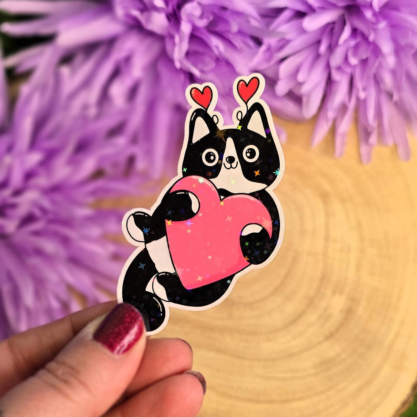 Sticker of a black and white cat holding a pink heart in front of purple flowers.