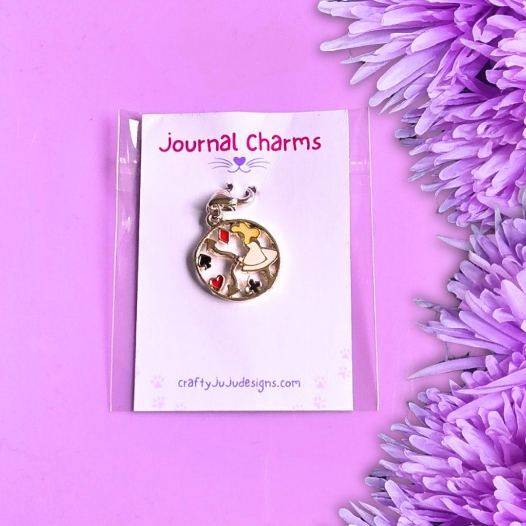 A round clock-shaped journal charm with a silver-toned metal frame and colorful details, displayed on a white card labeled 'Journal Charms' by Crafty Ju Ju Designs, set against a purple background with purple flowers on the side.