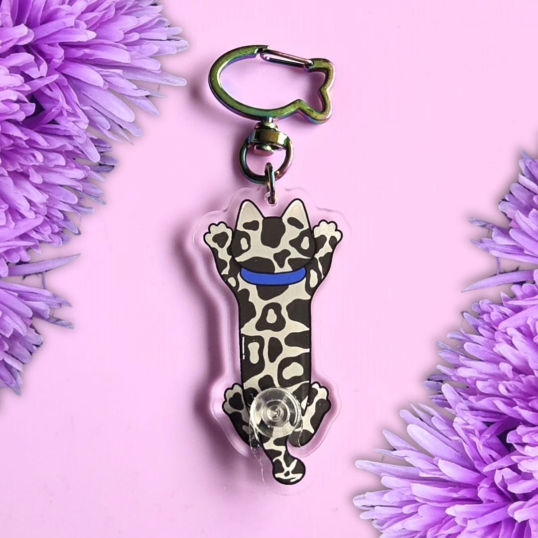 back view of grey tabby cat stretched out acrylic keyring with moving tail and fish shaped key ring clasp. All laid on a purple background with purple flowers around the edges