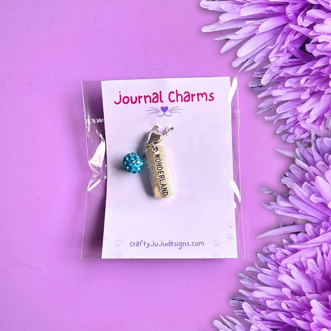 A small jewelry charm set in clear plastic packaging on a white display card labeled 'Journal Charms.' The set includes a silver-toned charm with 'WONDERLAND' printed on it and a blue beaded ball charm.