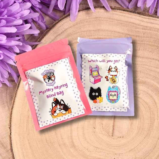 Two small colourful mystery keyring bags placed on a round wood slice, with purple flowers around the top edge and green grass underneath. The pouches have cartoon graphics and text including 'Mystery Hiding Treat Bag' and 'What will you give you?'.