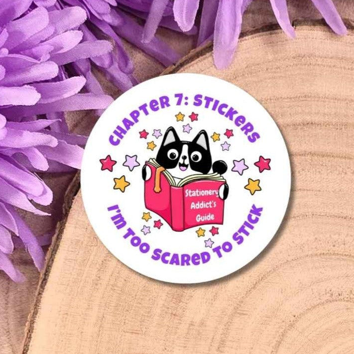 A round sticker featuring a black and white cat reading a pink book with purple text, displayed on a wooden slice with purple flowers.