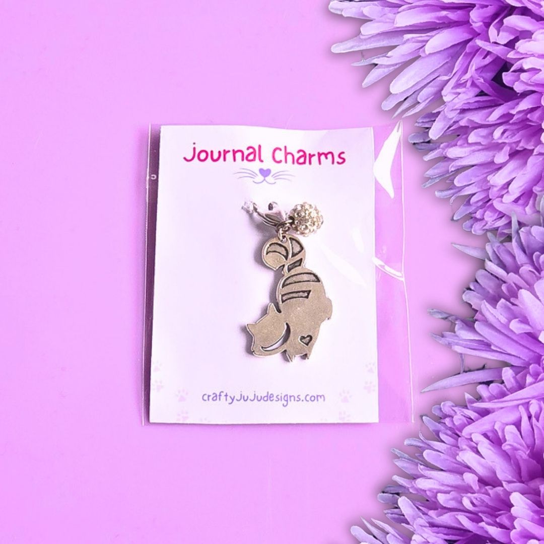 A cat-shaped journal charm displayed on a white backing card labeled 'Journal Charms' with the website craftyjujudesigns.com, set against a purple background with purple flowers on the right.