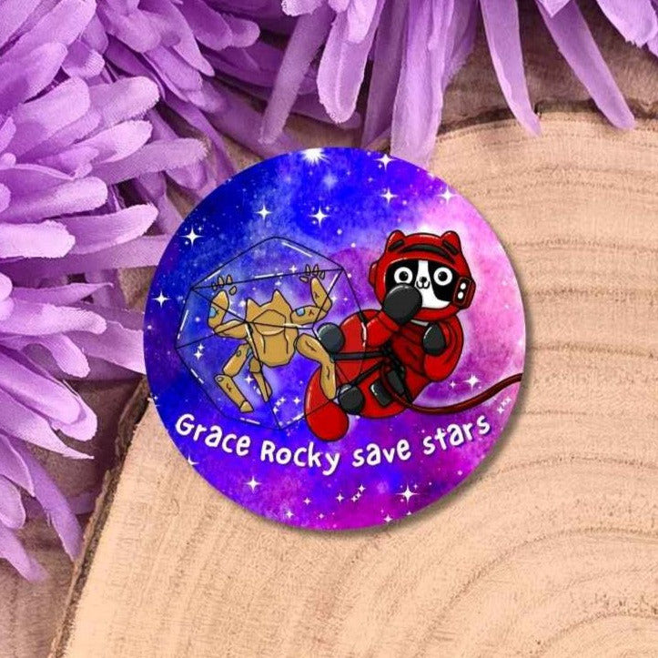 A round pin badge featuring a colourful space-themed illustration with a red cat in a space suit and a cat rocky, surrounded by purple stars, placed on a wooden surface with purple flowers in the background.
