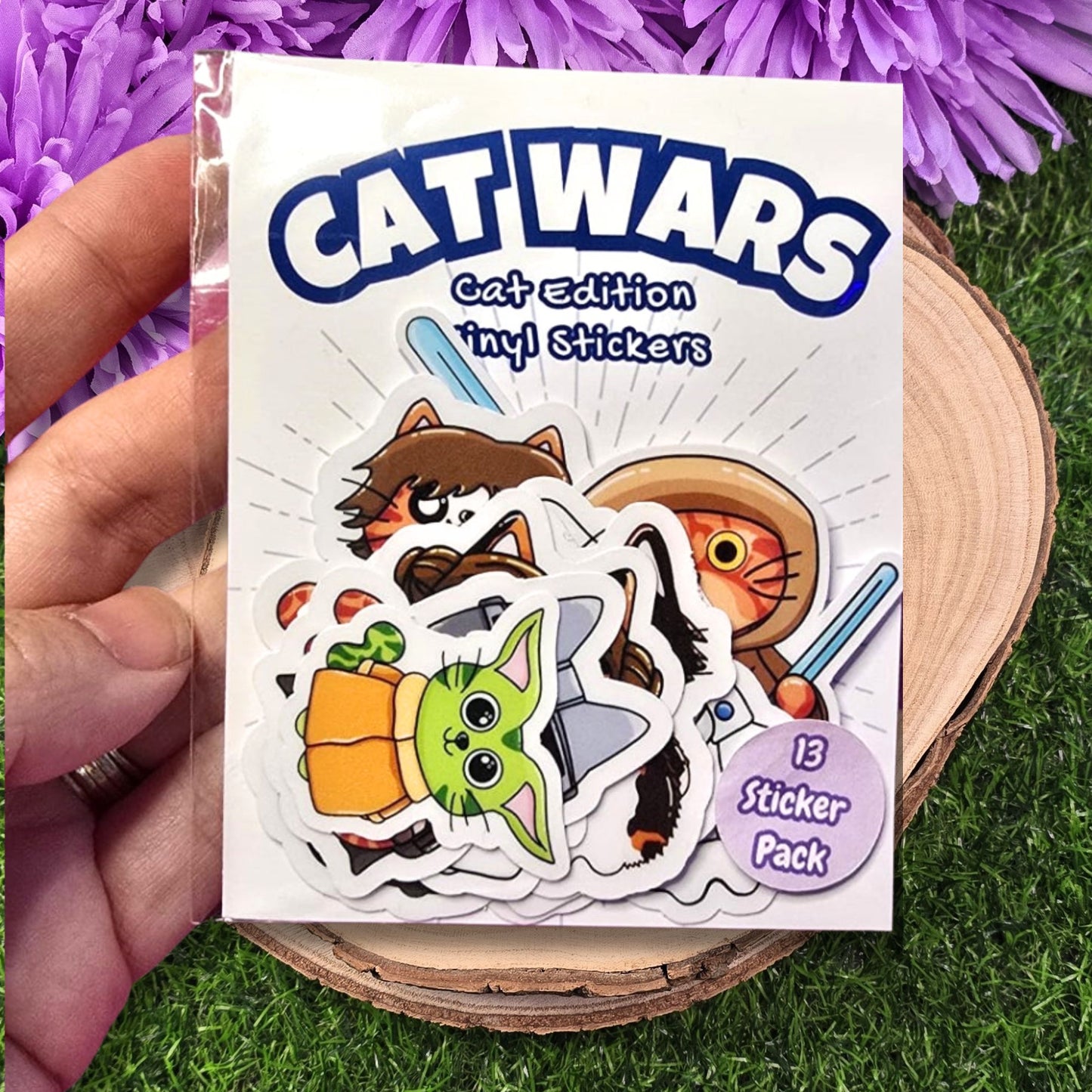 Cat-themed sticker pack with 'Cat Wars' branding held over a grass and purple flower background.